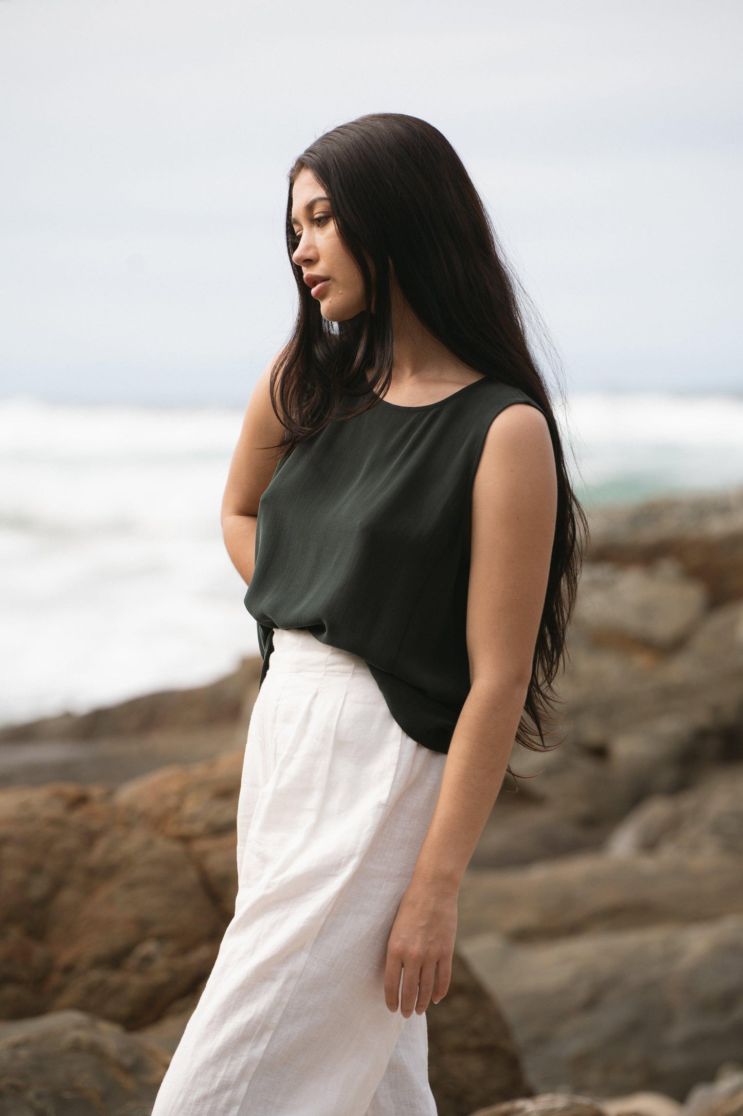 LILLY PILLY woman standing by the rocks, wearing a green LULU silk tank top and white OLI linen pants.