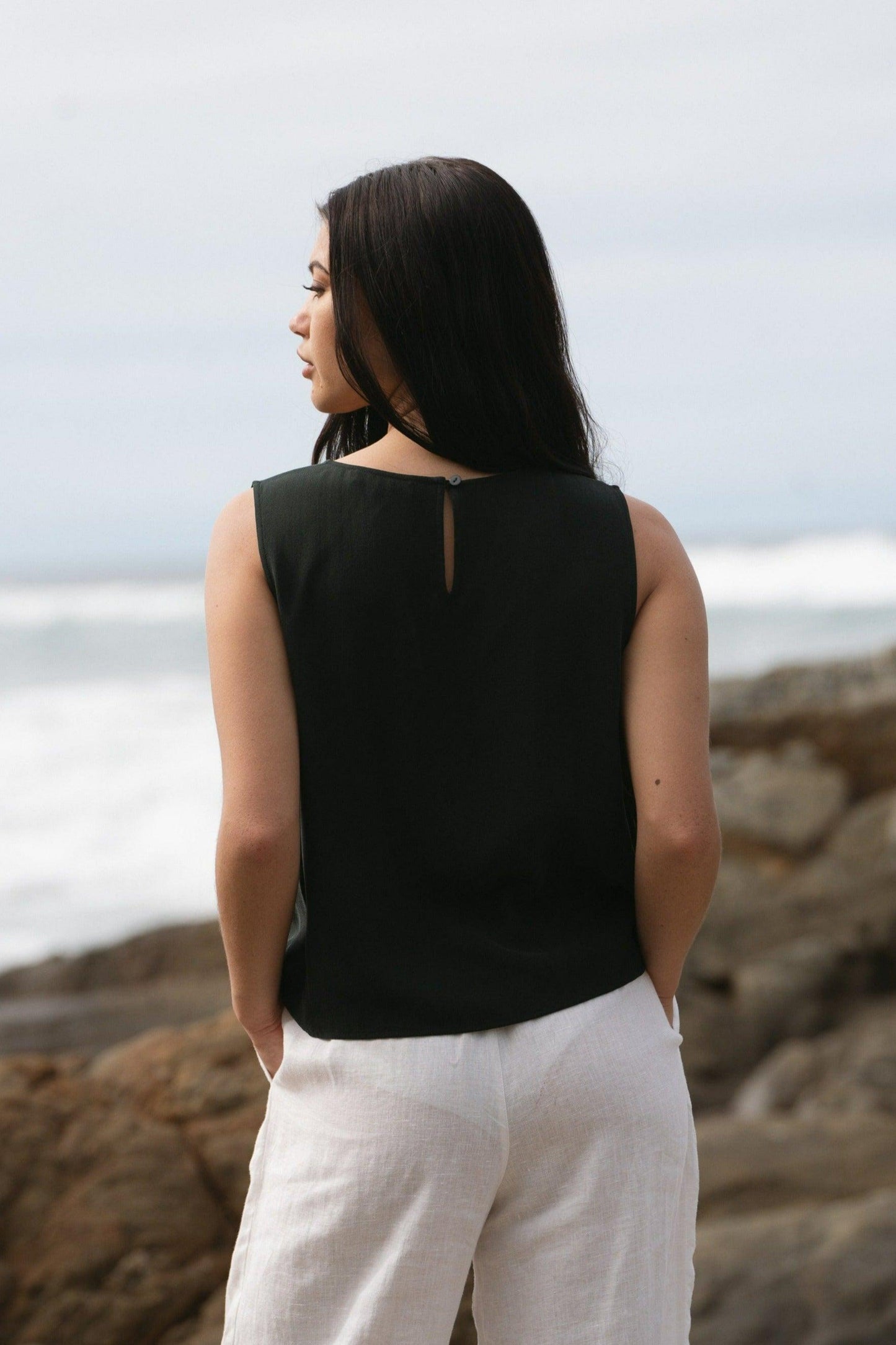 LILLY PILLY woman standing nearby the ocean, wearing a green LULU silk tank top and white OLI linen pants.