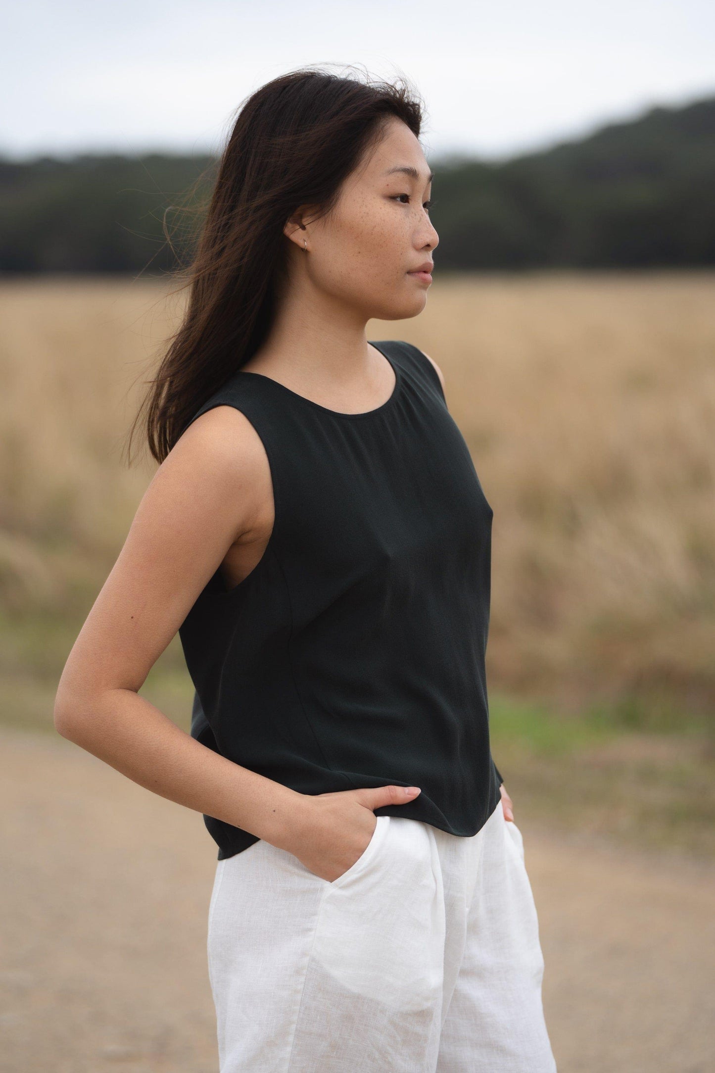 LILLY PILLY woman standing near a field, wearing a green LULU silk tank top and white OLI linen pants.