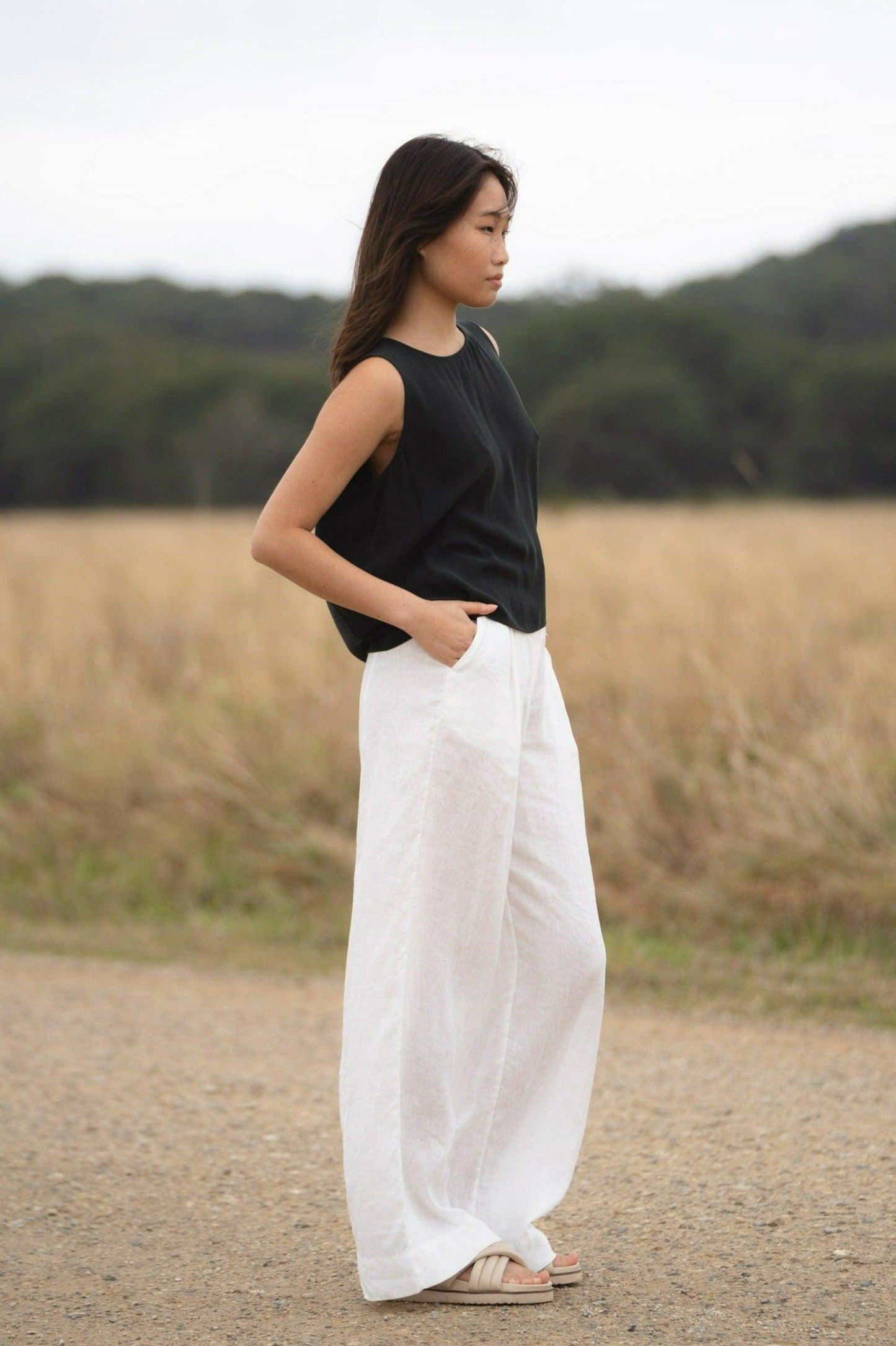 LILLY PILLY woman standing on a dirt road near a field, wearing a green LULU silk tank top and white OLI linen pants.