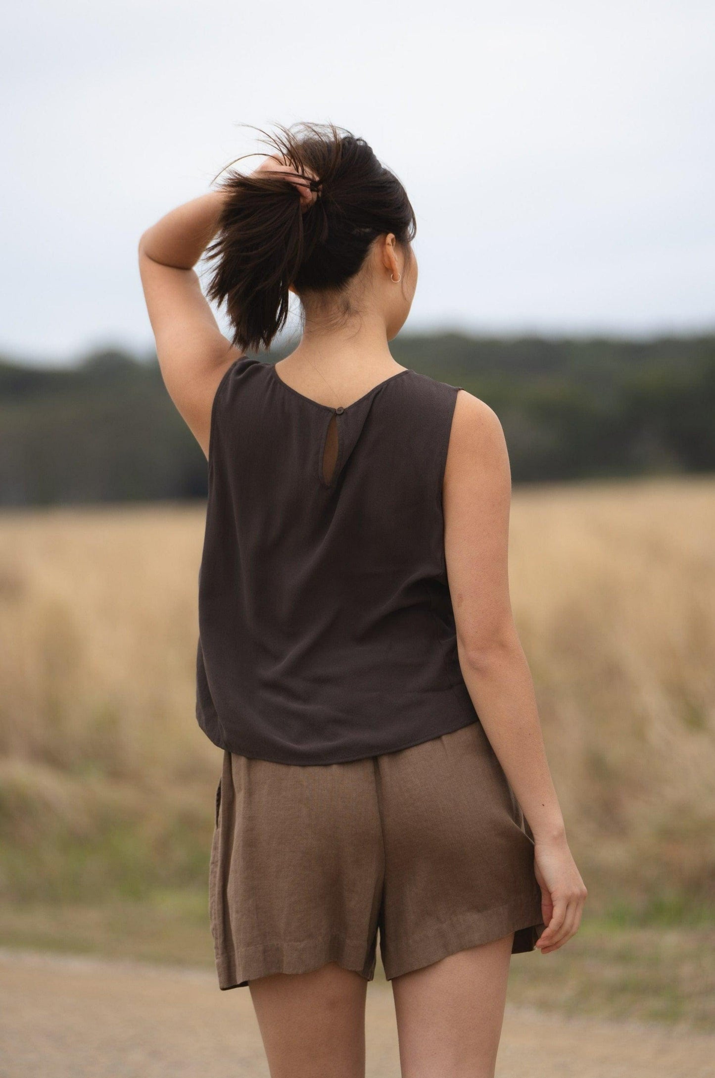 LILLY PILLY girl wearing a brown LULU silk tank top and DARK EARTH AMELIA linen shorts, standing on a dirt road with a field in the background.