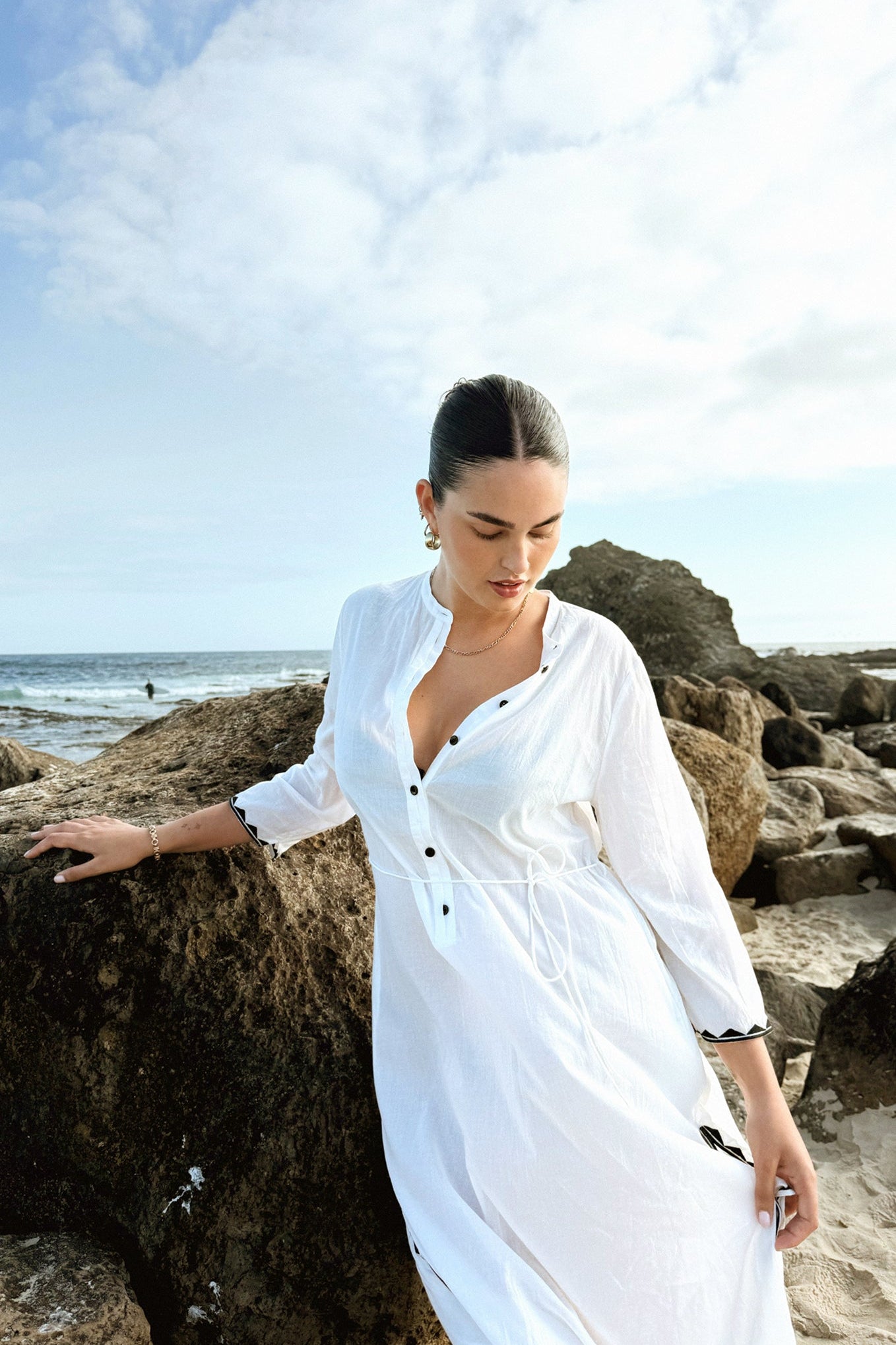 LILLY PILLY woman in a ivory Callie cotton dress standing on a rocky beach with ocean and sky in the background