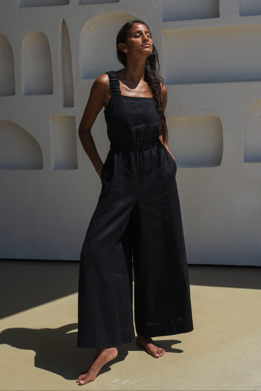 LILLY PILLY woman wearing a black Keely linen jumpsuit standing against a geometric wall.
