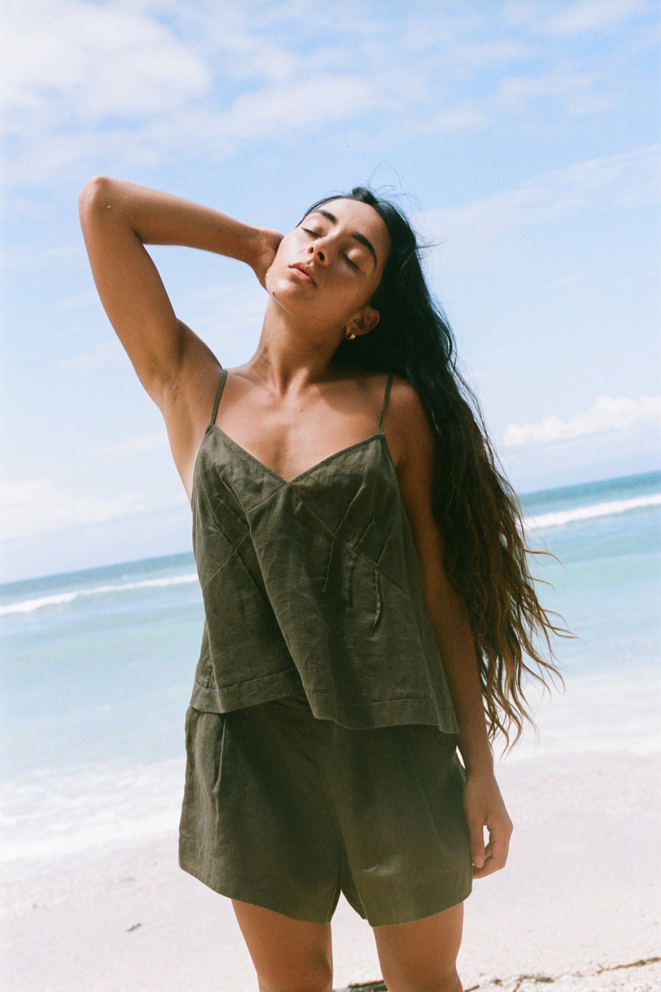 LILLY PILLY woman wearing a khaki Bea linen cami top and Gaia shorts standing on a beach with blue sky and ocean in the background