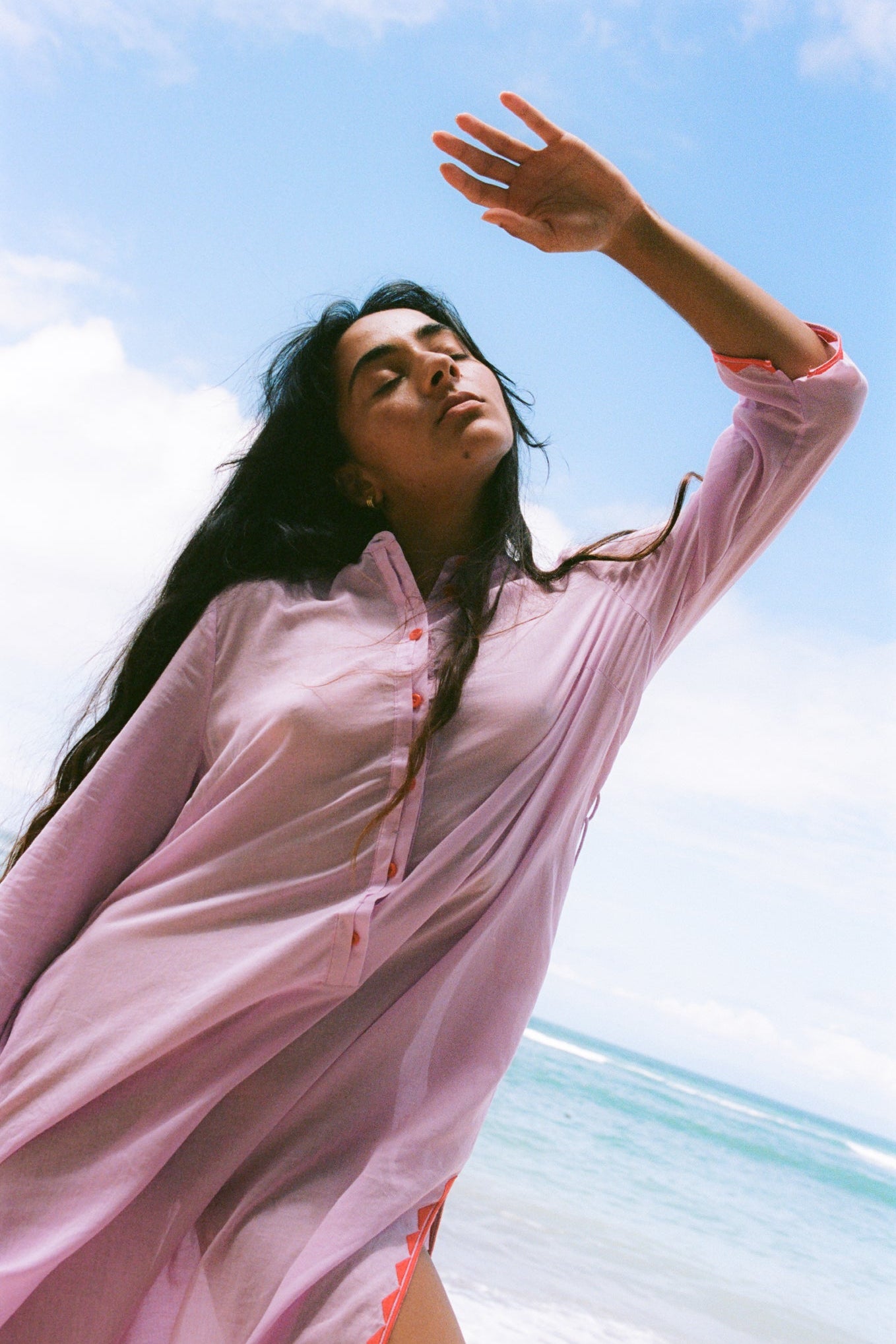 LILLY PILLY woman in a orchid Callie cotton dress standing on a beach with a clear blue sky.