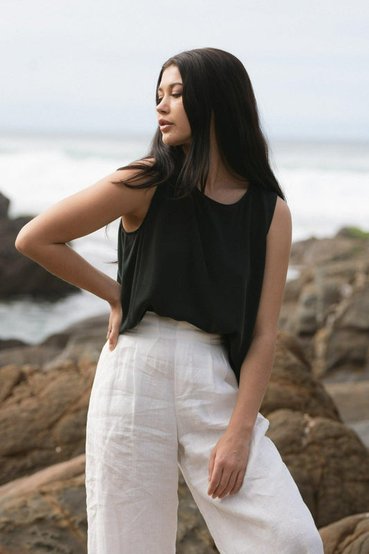 LILLY PILLY woman standing by a body of water, wearing a green LULU silk tank top and white OLI linen pants.