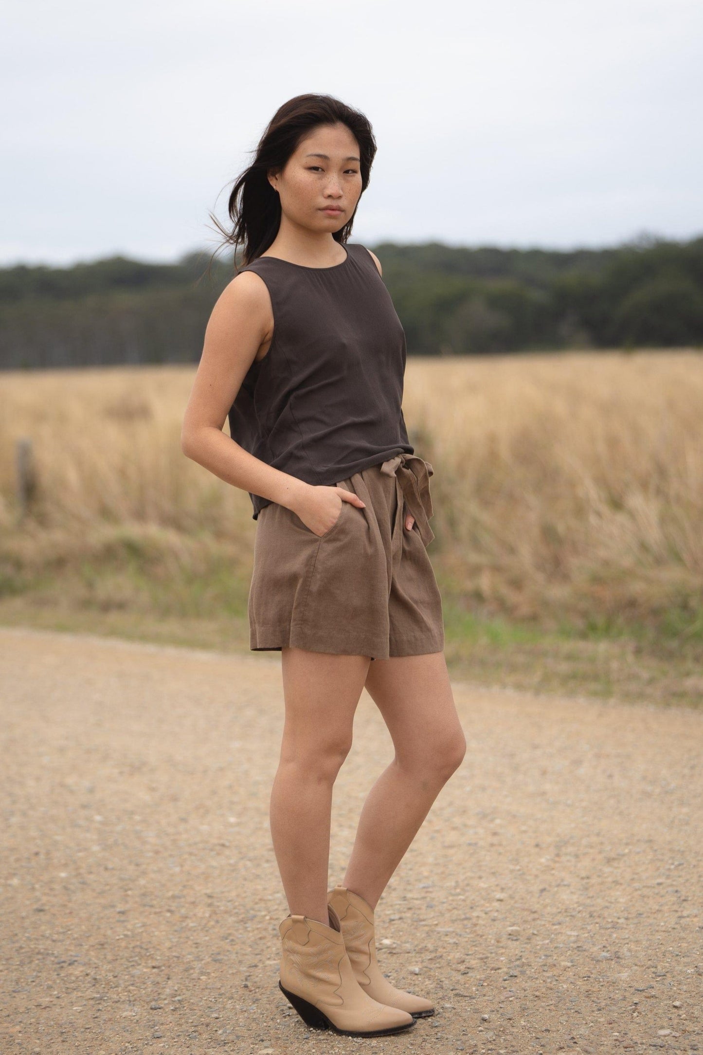 LILLY PILLY girl wearing a brown LULU  silk tank top and DARK  EARTH AMELIA linen shorts, standing on a dirt road