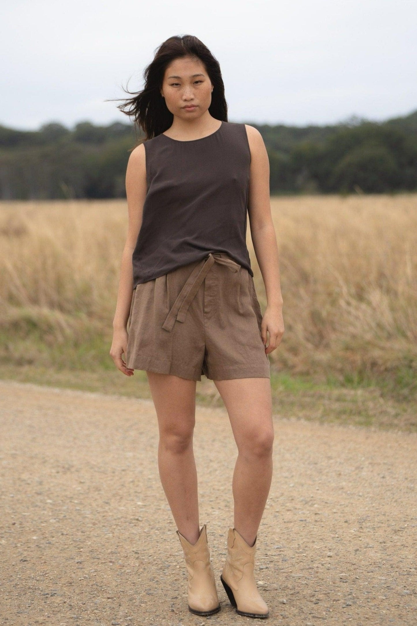 LILLY PILLY girl wearing a brown LULU  silk tank top and DARK  EARTH AMELIA linen shorts, standing on a dirt road with a field in the background.