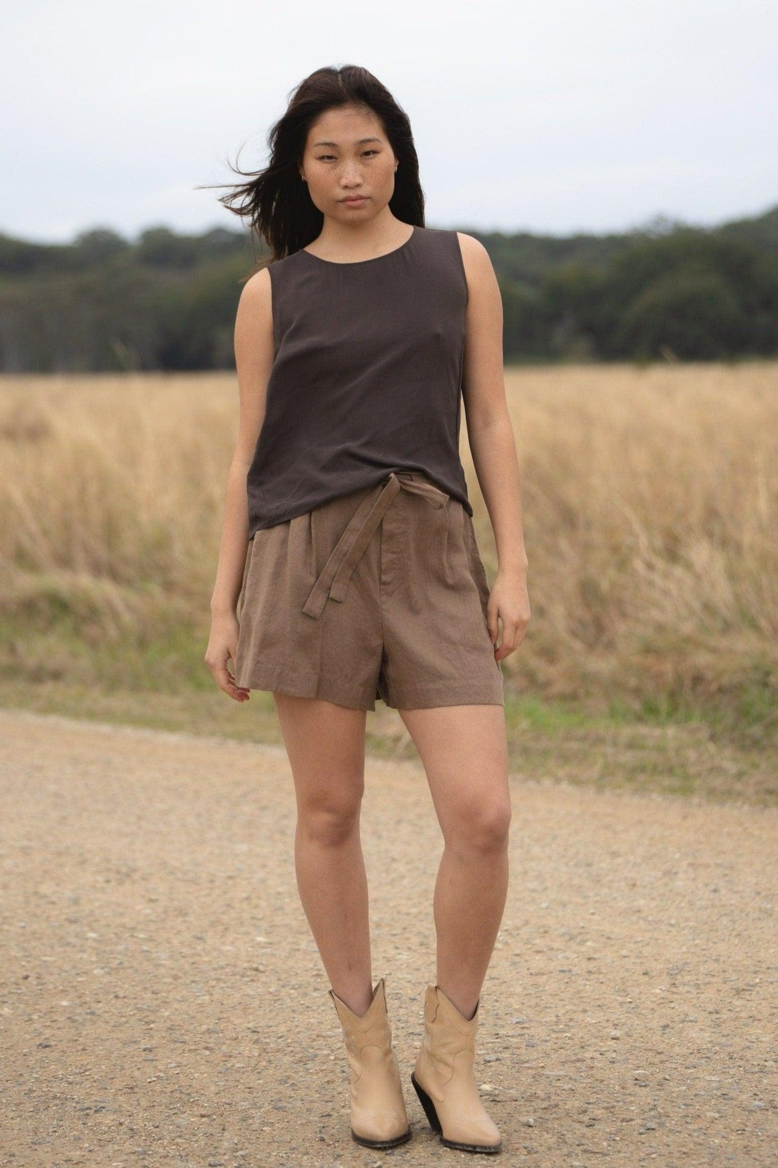 LILLY PILLY girl wearing a brown LULU  silk tank top and DARK  EARTH AMELIA linen shorts, standing on a dirt road with a field in the background.