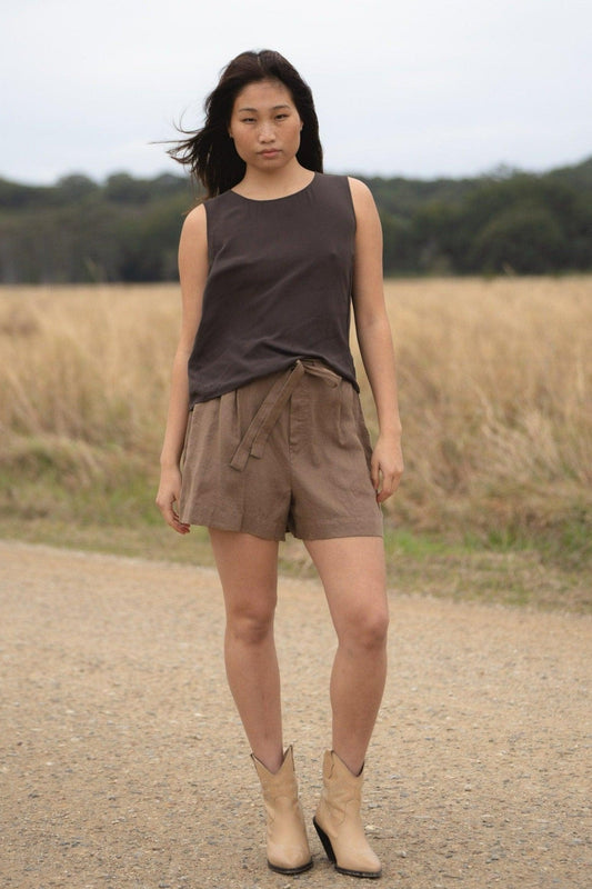 LILLY PILLY girl wearing a brown LULU  silk tank top and DARK  EARTH AMELIA linen shorts, standing on a dirt road with a field in the background.