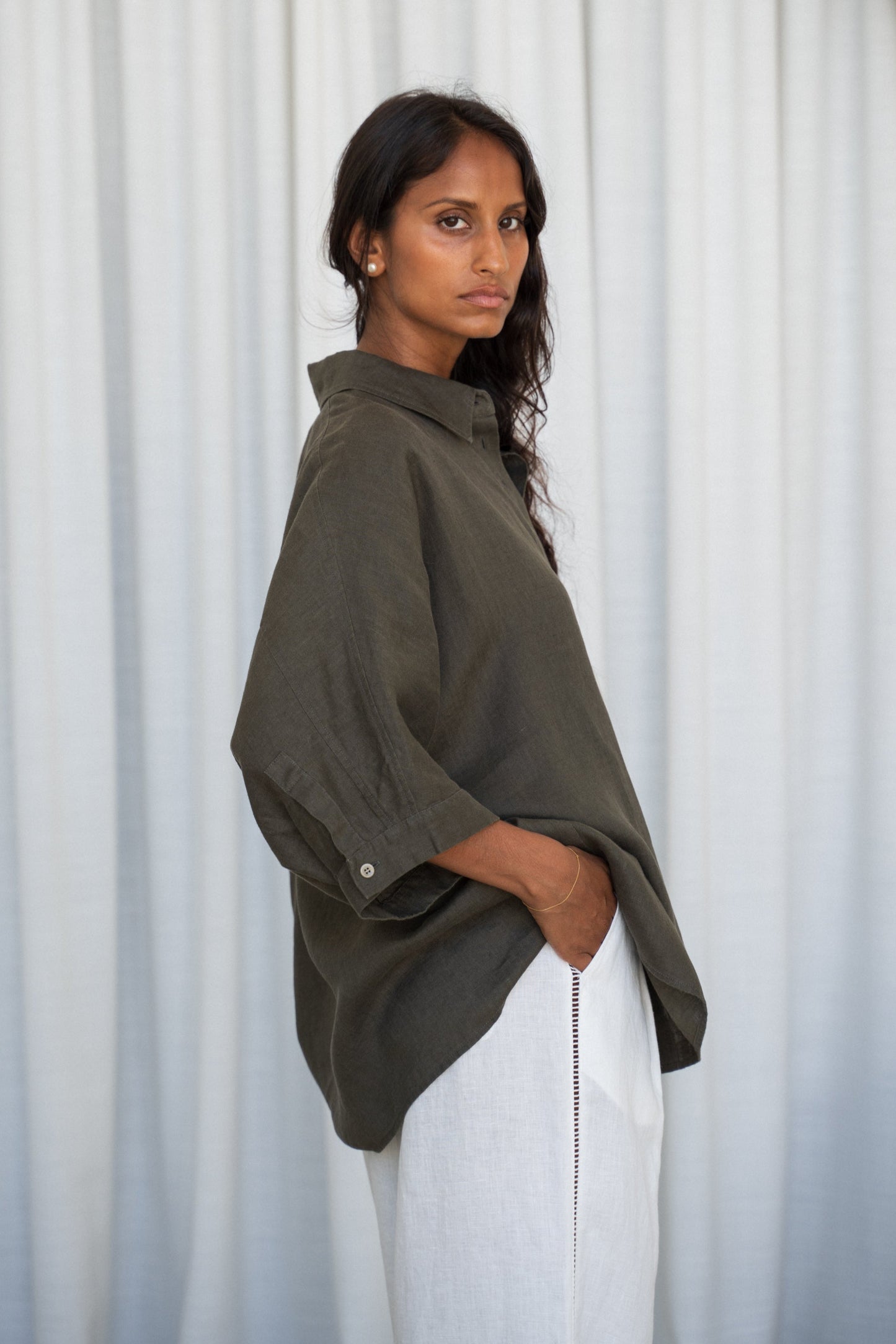 LILLY PILLY woman side view wearing a khaki Pixie linen shirt and Oli white linen pants outfit against a white curtain background