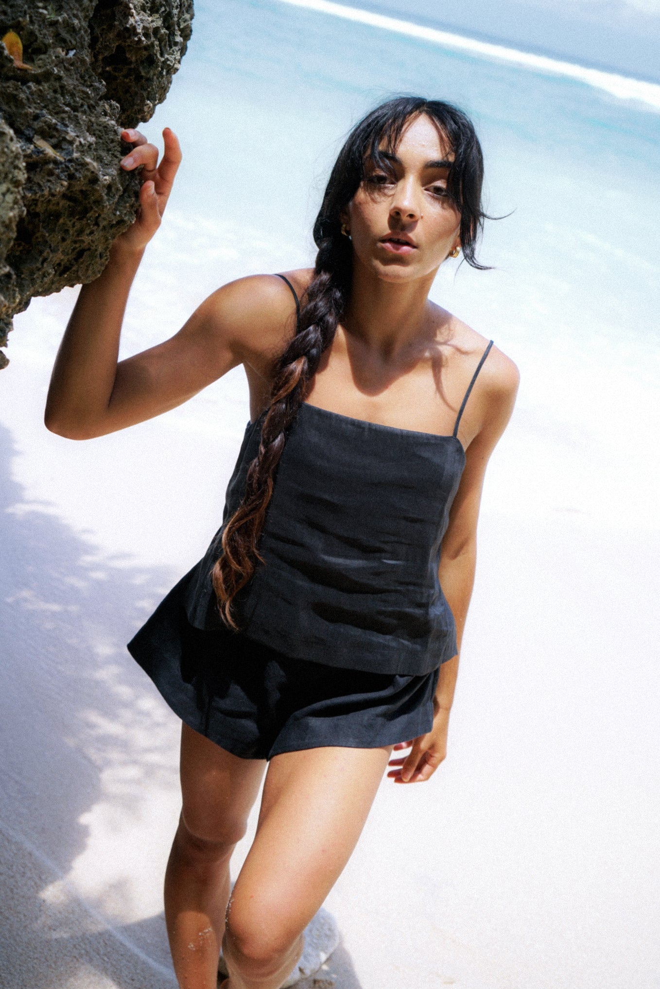 LILLY PILLY woman wearing a black Rosa silk linen cami top and shorts standing by a rock on a beach