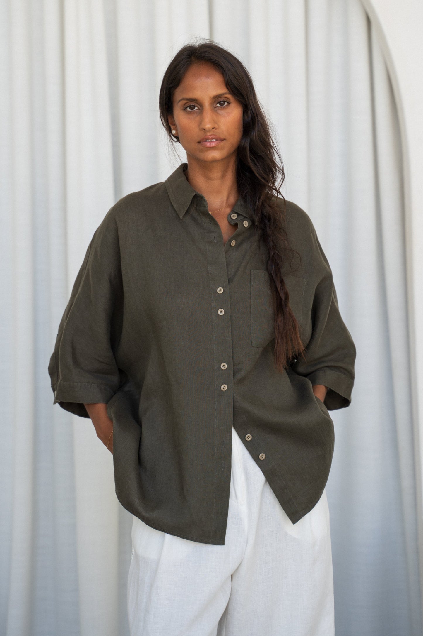 LILLY PILLY woman wearing a khaki Pixie linen shirt and Oli white linen pants outfit against a white curtain background